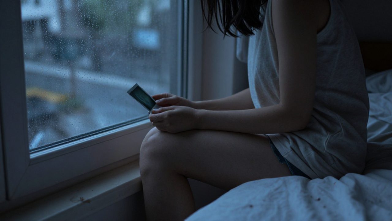 Persona sentada en una cama mirando por la ventana lluviosa mientras sostiene una fotografía antigua.