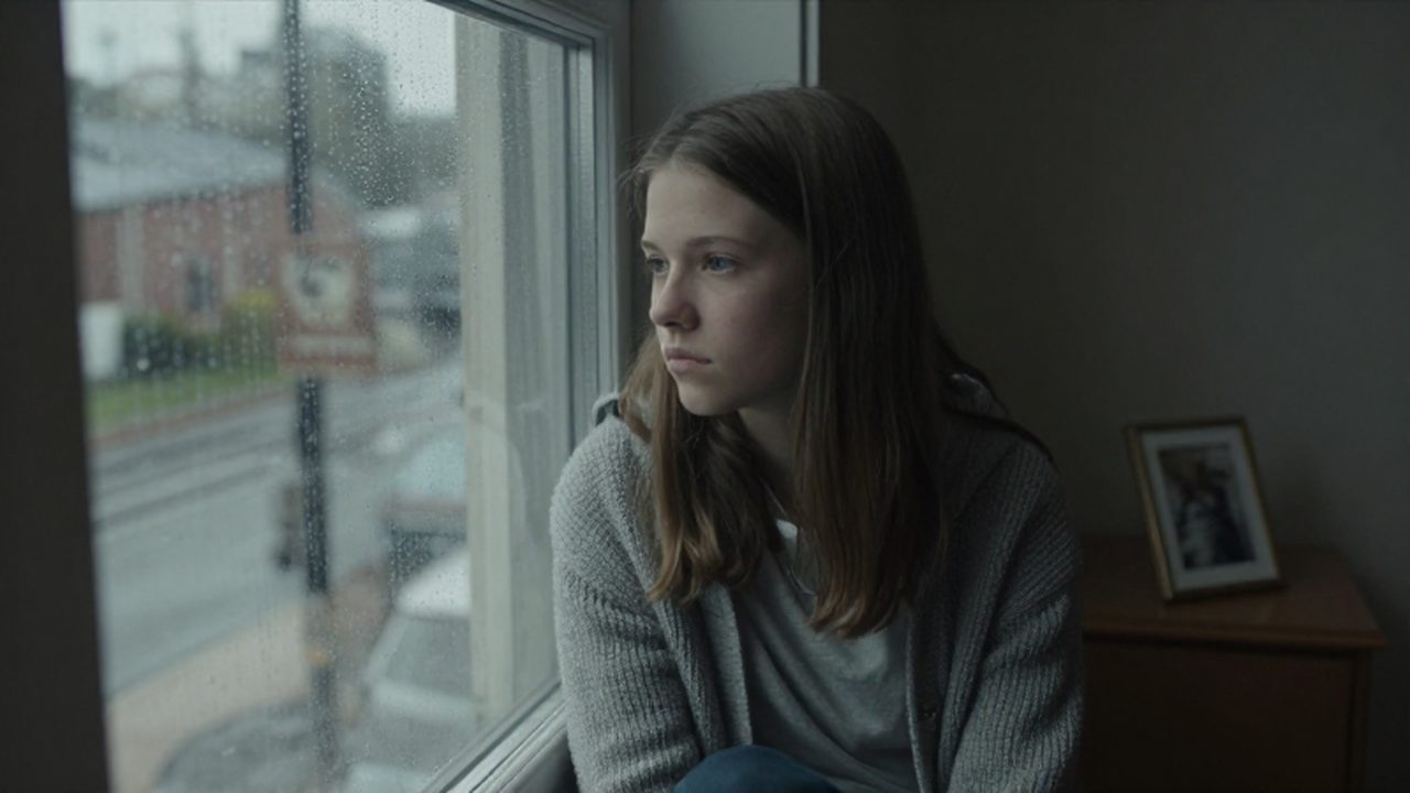 Una adolescente con expresión melancólica observa la lluvia desde el asiento de una ventana.