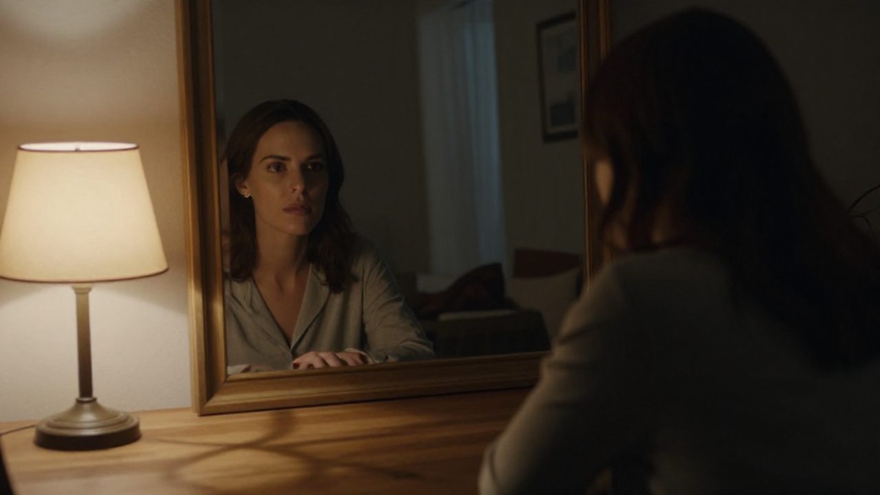 Mujer sentada a la mesa de madera contemplando su reflejo borroso en un espejo.
