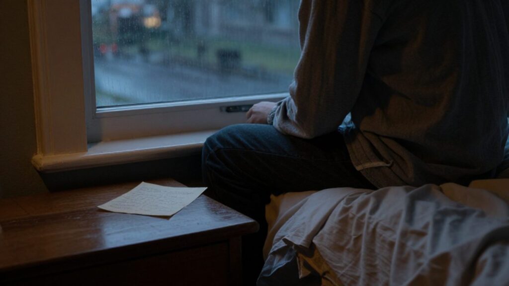 Persona solitaria sentada en el borde de la cama mirando hacia una ventana lluviosa durante el crepúsculo.
